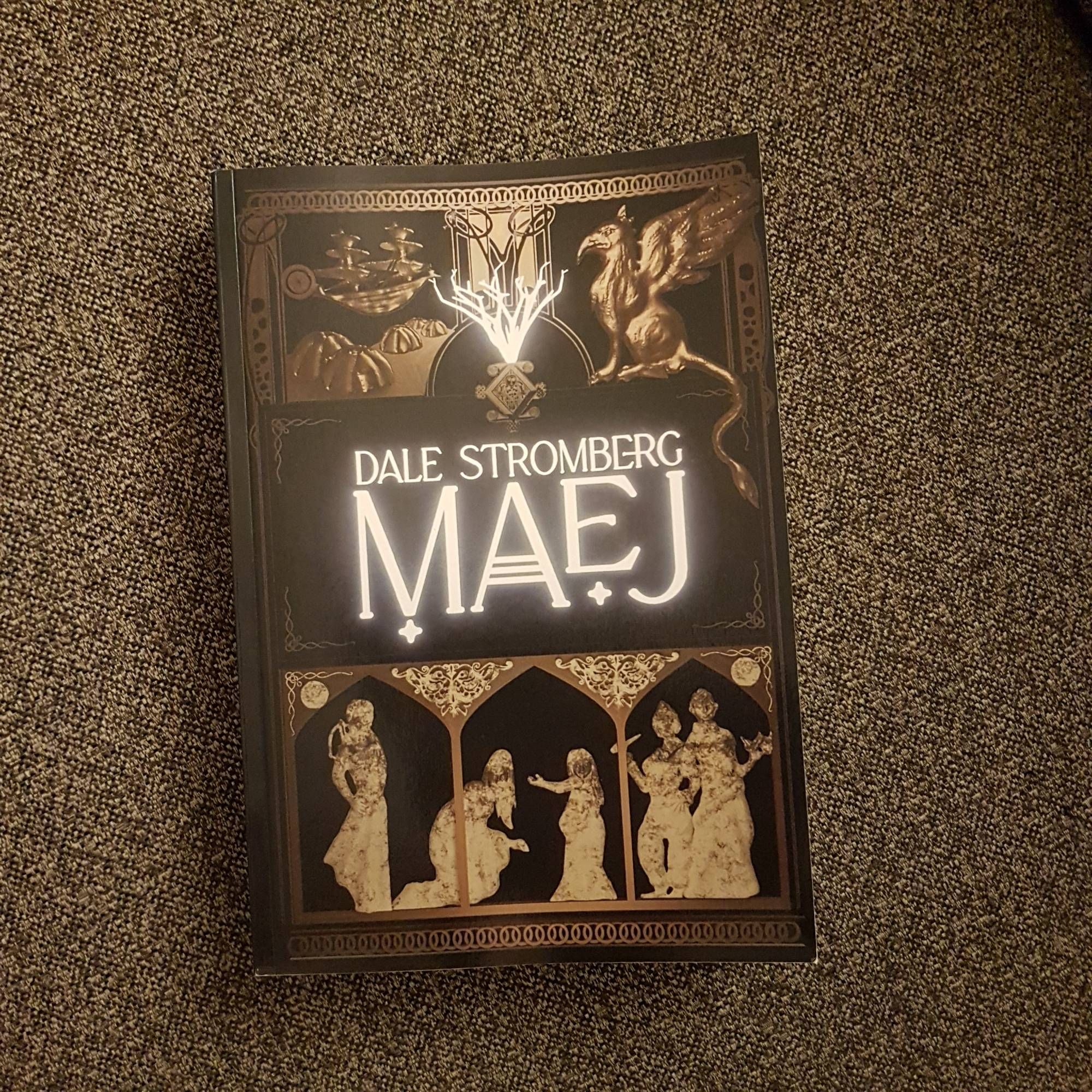 A picture of a physical copy of MAEJ. Dark background, with feminine sculpted figures, gryphons and ships. Luminous letters say MAEJ and Dale Stromberg. Cover by Rachel A. Rosen.