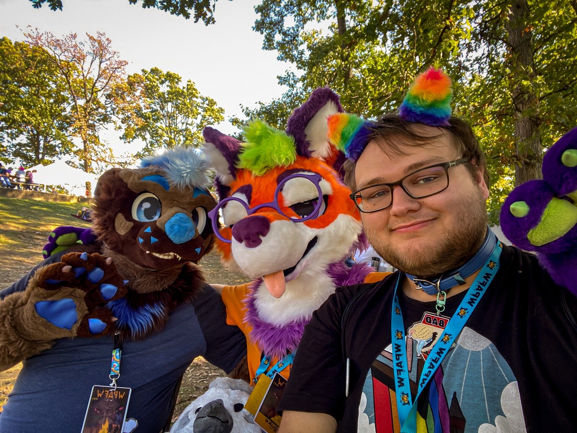 a group photo of two fursuiters (a brown-and-blue otter and a vibrant maned wolf) and a person wearing rainbow ears