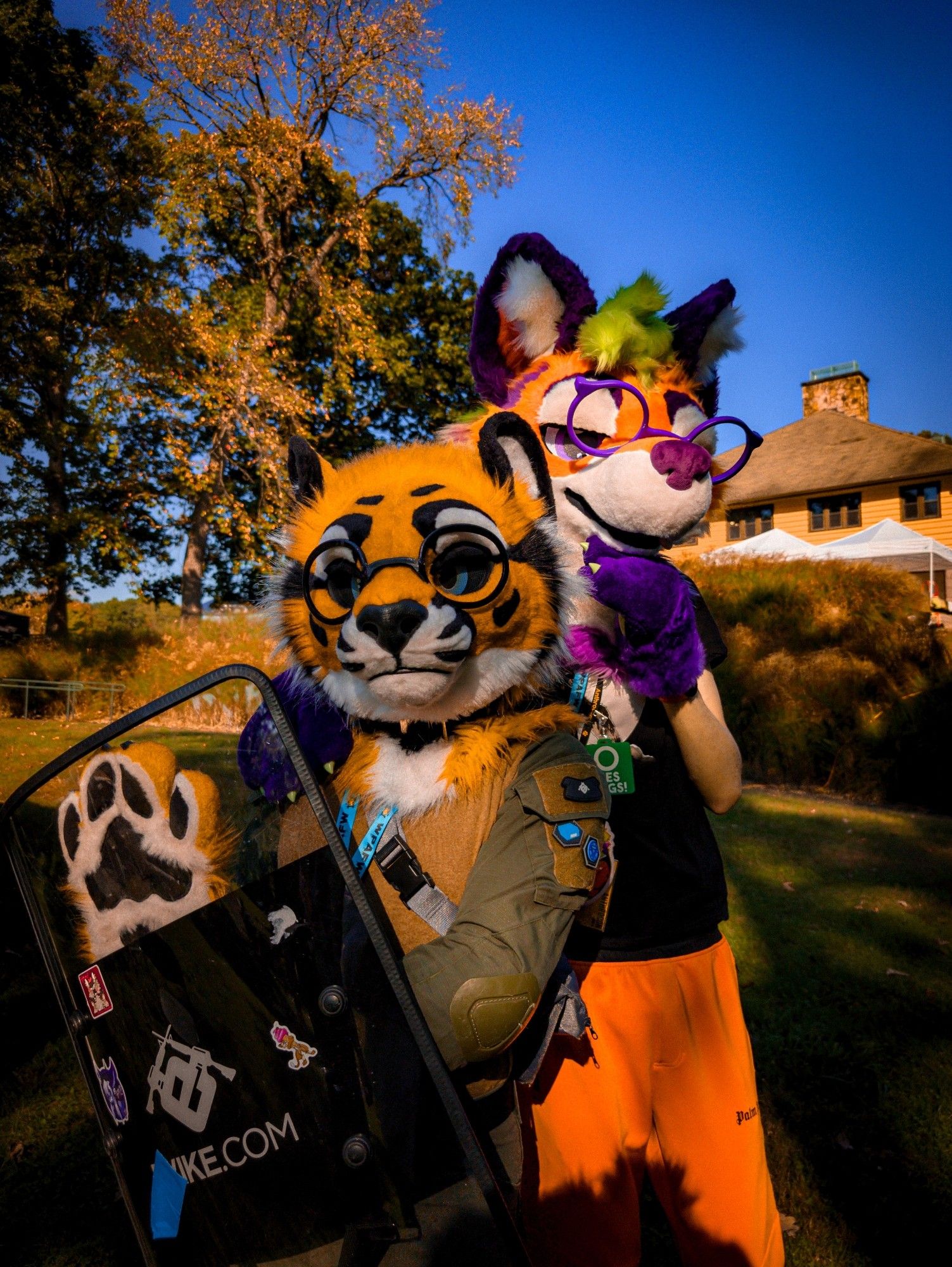 two fursuiters posing against a lodge in a park: a yellow-and-black in tactical gear is in the front holding a shield; a vibrant maned wolf stand in the back with his head resting on one of the paws