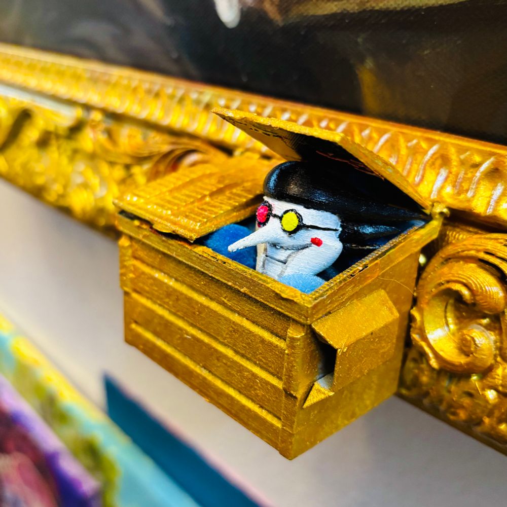 A close-up of the bottom edge of the opulant gold frame shows a gold 3d printed dumpster.  Spamton, a trash-dweller, peaks his beautiful, horrible head out from under the lid.  The dumpster around him is brimming with blue spheres, known as pipis.