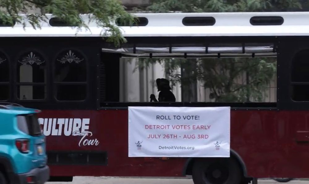 A bus passes with the silhouette of a passenger inside. A sign on the side of the bus states," Roll to vote1 Detroit votes early. July 26th - Aug. 3rd