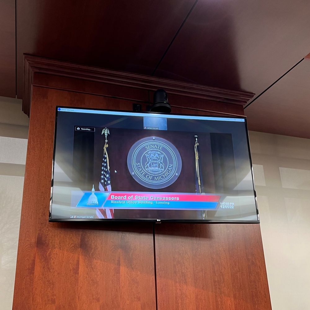 A monitor on a wall shows a livestream of a previous Board of State Canvassers meeting