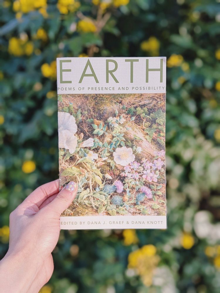 My left hand holding a copy of the Earth anthology in front of a green bush with yellow flowers.