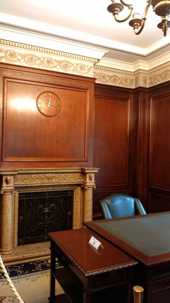 クラシックな執務室の写真。重厚な暖炉の上の壁に時計。天井にシャンデリア。大きい机に革張りの椅子。
