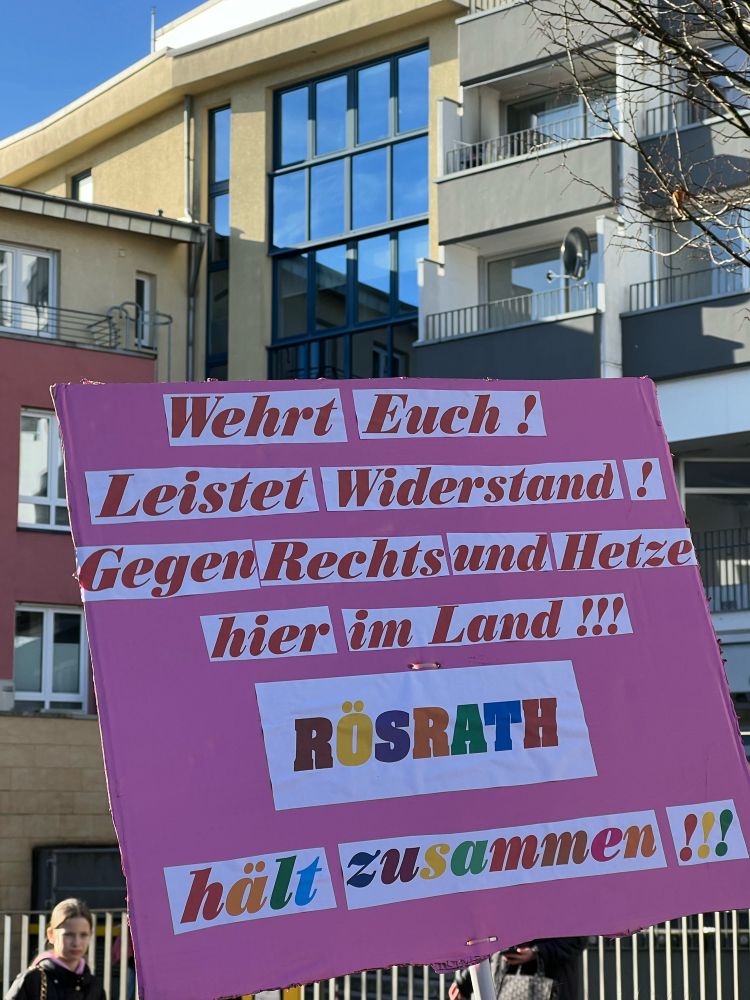 Protestplakat mit rosa Hintergrund. Darauf steht in rot auf weiß „Wehrt euch! Leistet Widerstand! Gegen Rechts und Hetze hier im Land!!!“ In bunten Buchstaben auf weiß steht darunter „Rösrath hält zusammen!!!“