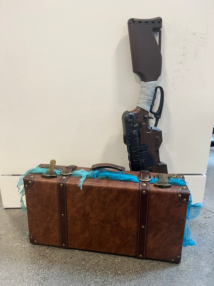 a brown vintage-looking suitcase with a frayed neon blue/green scarf peeking out the edges, simulating magical auras. behind it is a nerf slingfire, painted to look more like an actual lever-action gun