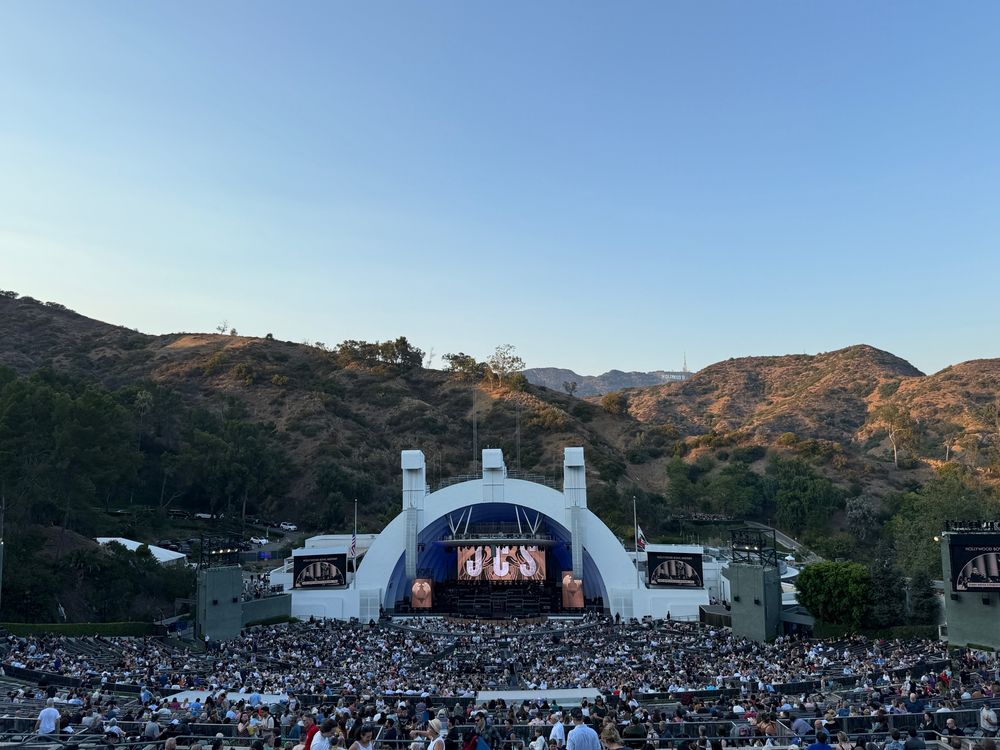 View of the half-filled the Hollywood Bowl from the back of the auditorium, the Hollywood sign in the background. Screens on stage are showing JCS.