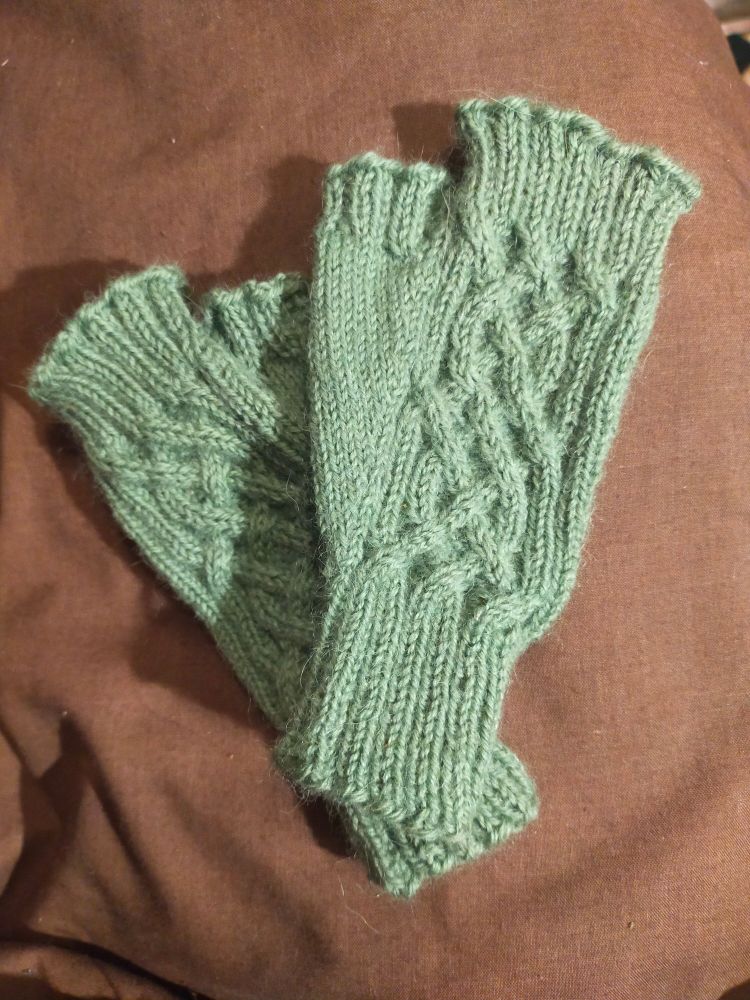 Two light green knitted mitts on a brown background. The mitts have a cable design on the back of the hand.
