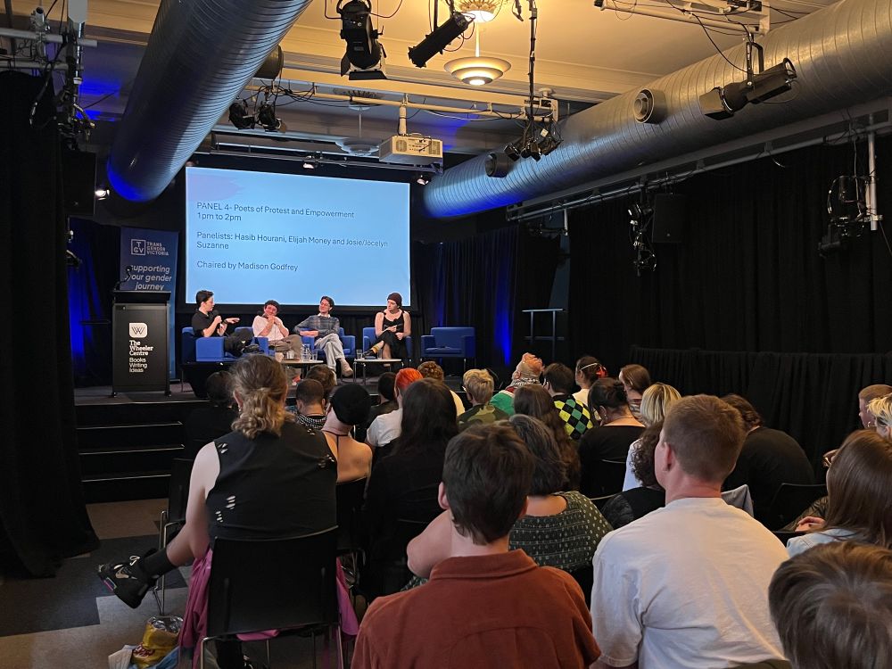 The back of the image contains a stage with four people sitting on blue chairs. The backs of audience members are in the foreground of the image. 