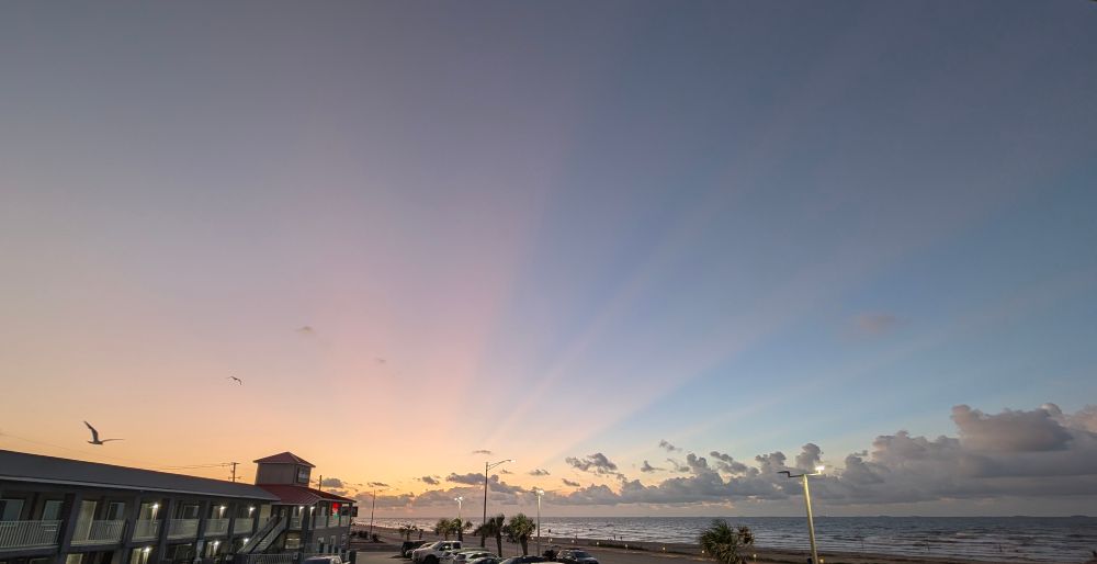 Shafts of light streak across a blue morning sky as the sun rises over the beach in Galveston Texas.