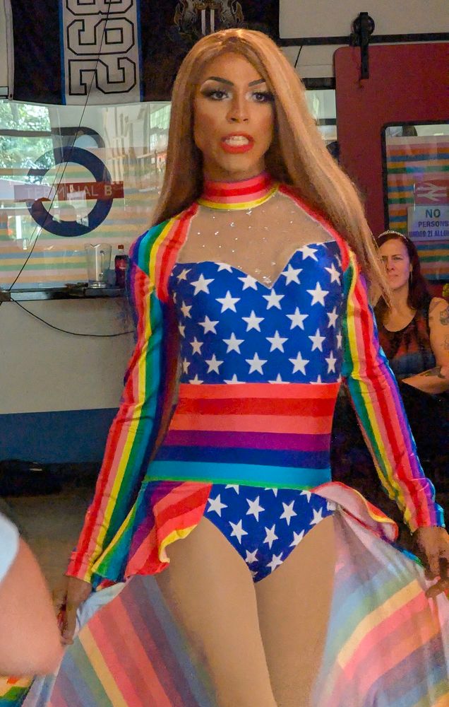Kiki Diamond struts in a blue bodysuit with white stars, topped by a rainbow sash, train, and sleeves