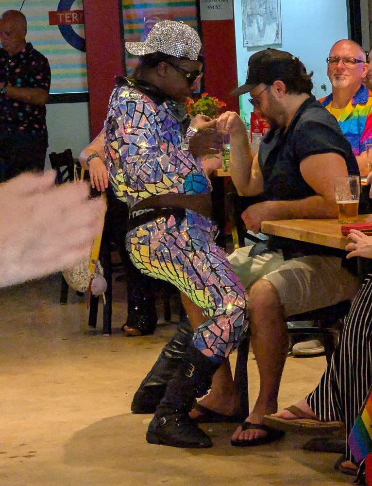 A dancer in an outfit that looks like it's made of broken glass dances with a man sitting at a table.