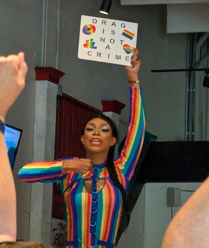 In a rainbow-striped outfit, Kiki Diamond holds up a sign that reads, "Drag Is Not a Crime"