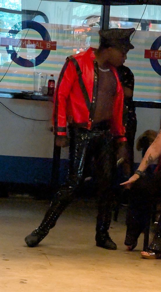 A performer in a vibrant red jacket with black details, shiny black pants and a biker hat.