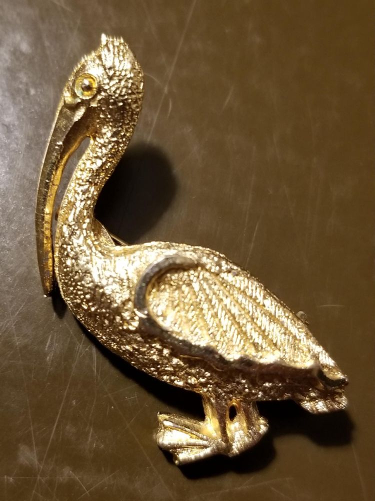 Gold-colored metal pin of a pelican standing upright with its bill held down along its chest.