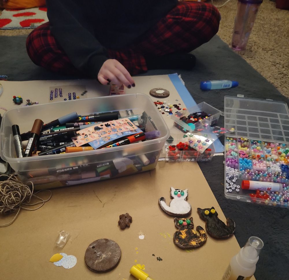 me & sib making Christmas ornaments on the floor. There is a bucket of paint markers, a tray of beads, pompoms, sequins, and other craft items. 