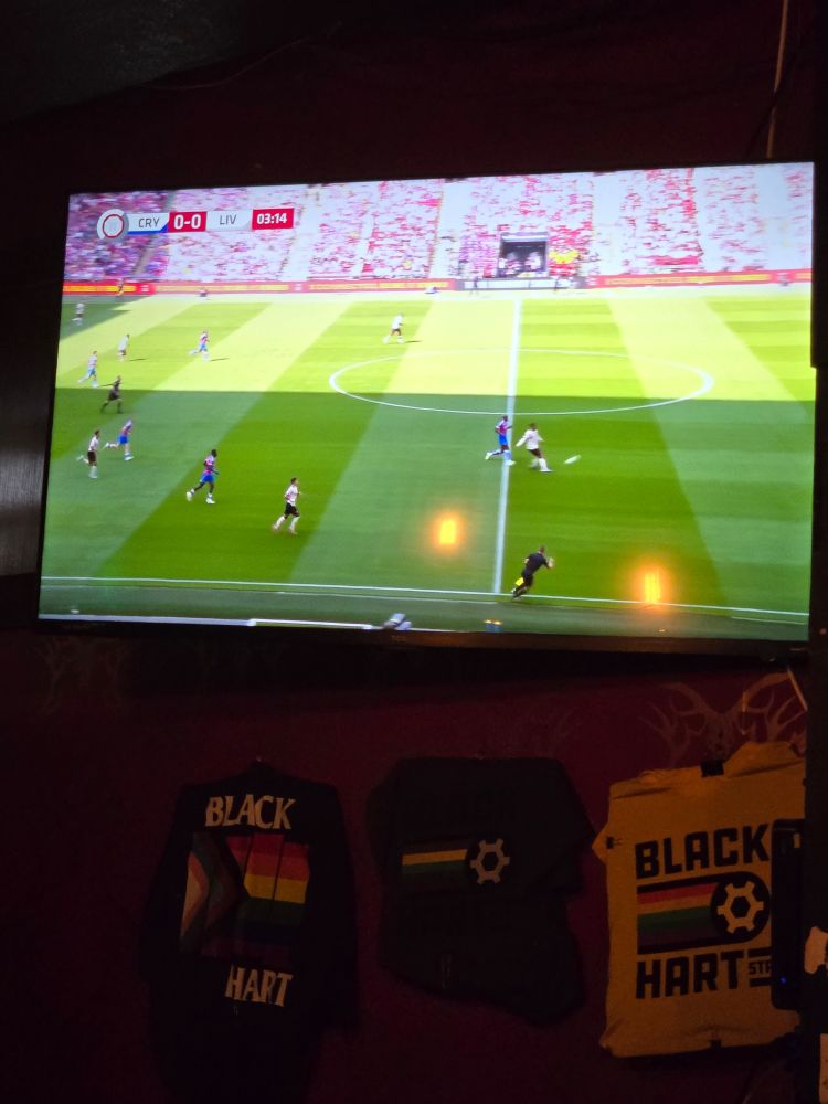 A TV screen showing a football match between Liverpool and Crystal Palace. Below the TV on the wall are hanging some Black Hart t-shirts.