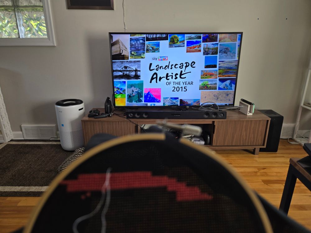 In the background is a TV with an advertisement for the BBC show Landscape Artist of the Year. Blurry, in the foreground, is a cross stitch hoop with a scattering of red stitches. 