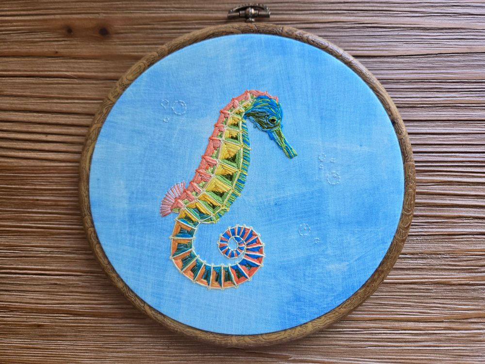 An embroidery hoop with blue fabric and a stitched seahorse. The seahorse has a blue and green head and belly, a yellow and green side, and an orange and green back. The spiraled tail is a blend of orange and blue