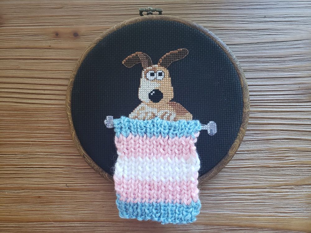 An embroidery hoop on a wooden background. The hoop has black fabric and the cross stitched cartoon character 'Gromit' from Wallace and Gromit. Gromit is holding knitting needles with knit fabric in the trans colors hanging from them. 