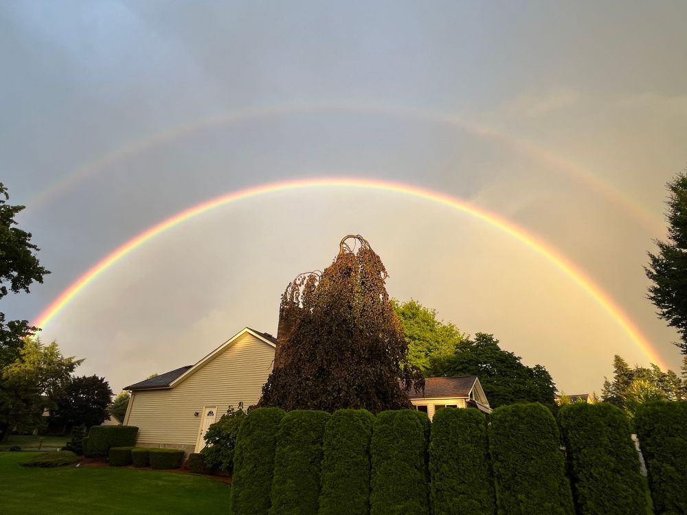 The arcs of a double rainbow stretching over a light colored house and dark green tree. The sky is cloudy but bright beneath the inner rainbow and darker above the outer rainbow 