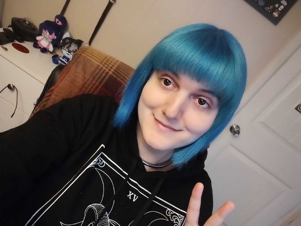 Roxie sits in her desk chair wearing a black hoodie with THE DEVIL tarot card image screen printed on it. Her hair is a vibrant blue from being freshly dyed and treated!!!