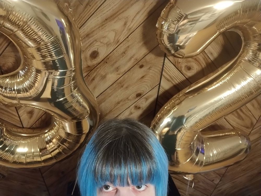 A giant pair of golden balloons in the shape of a 3 and a 2 float in the background with the top of a blue and black haired trans puppy gal peeking up from the bottom of the frame.