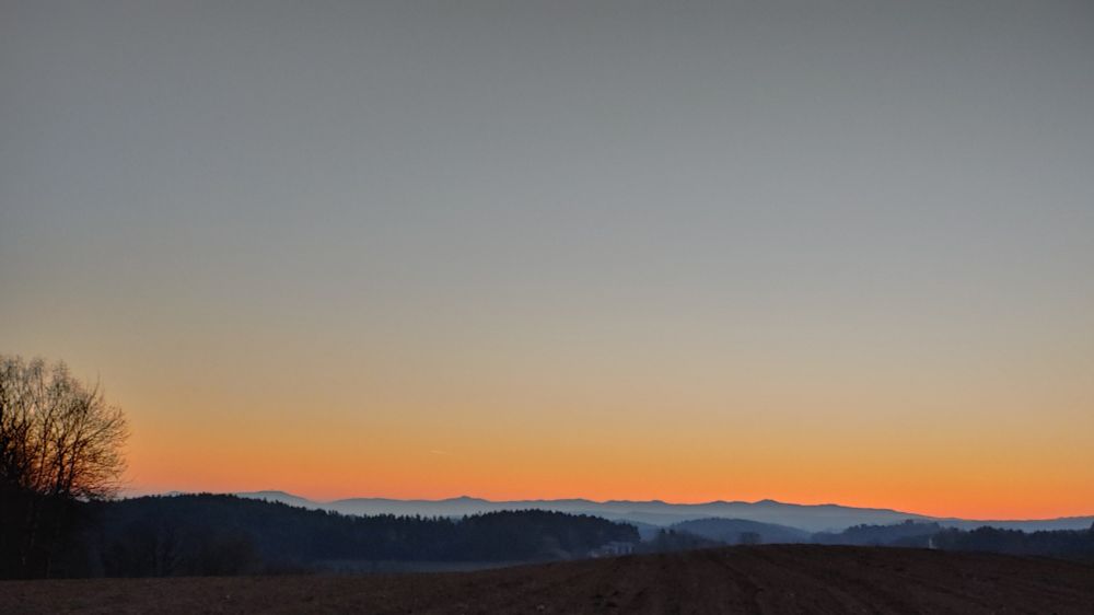 Silhouette of a landscape with soft blue hills and dark trees in the distance against a pale orange and blue morning sky. In the foreground a dark field and a leafless tree to the left.