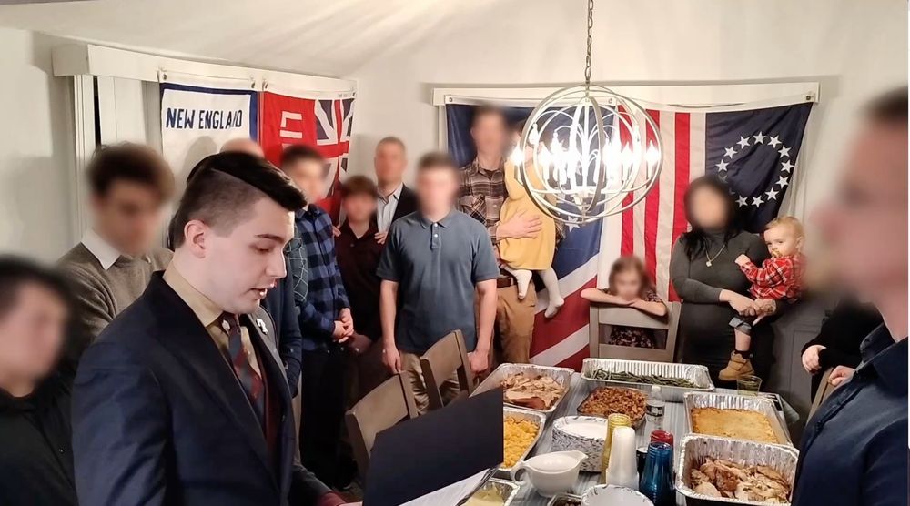 A still image from a video that shows a leader of white nationalist American Freedom Party giving a short speech to members of Patriot Front before digging into a holiday dinner. About 10 Patriot Front dorks, and a few of their children and partners stand around the dinner table that is full of food. Their faces are poorly blurred in an attempt to hide their identities, but it is possible to see who they are. The only one that doesn't not have a blurred face is the leader of American Freedom Party. 
