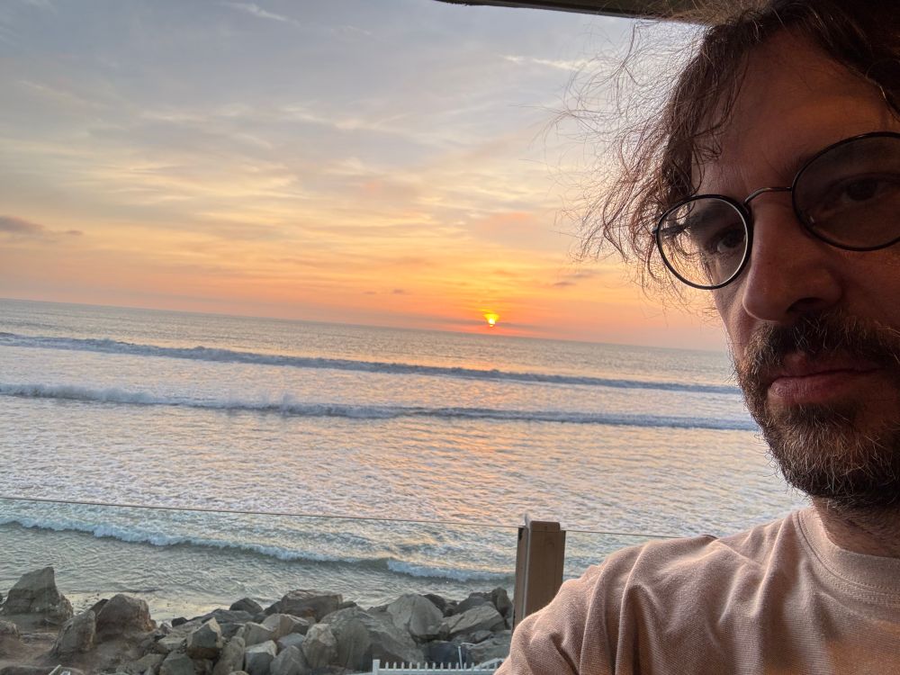standing behind a glass balcony overlooking rocky coast with a sun setting over the Pacific 