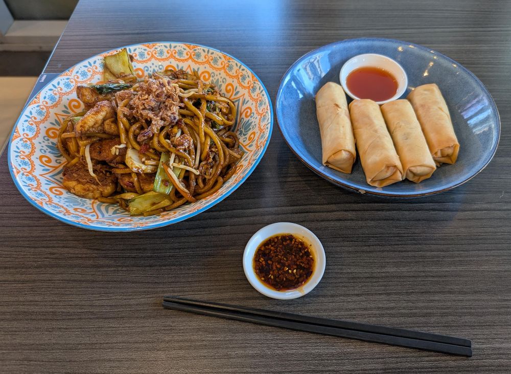 A plate of mie goreng next to a plate with four spring rolls. Chopsticks and a bowl of chili oil next to them.