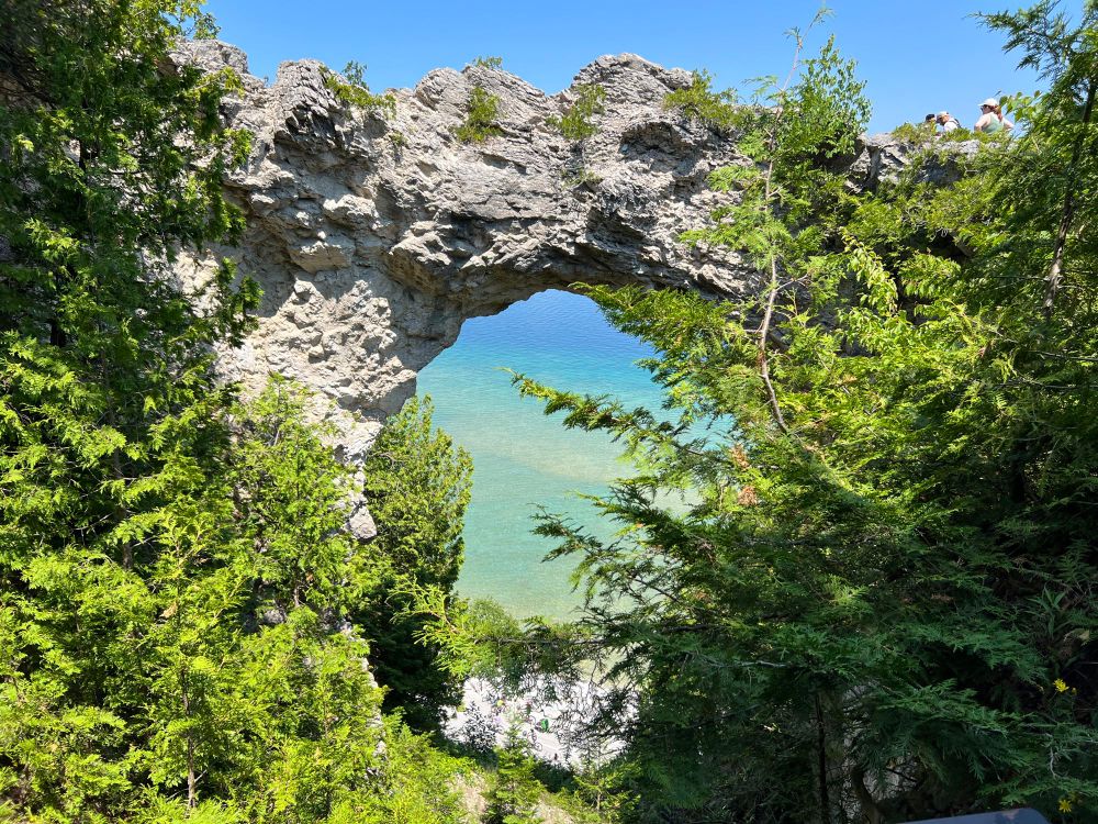 A natural limestone arch frames the crystal-clear turquoise waters below, surrounded by lush green foliage under a bright blue sky.
