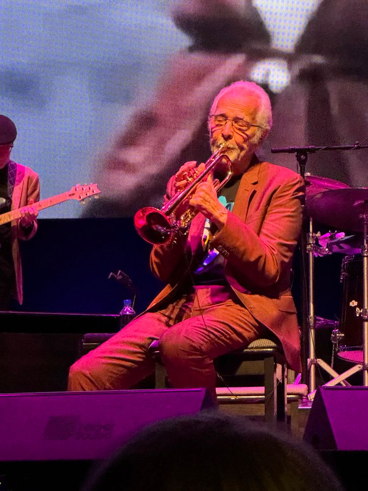 Close up of Herb Alpert sitting on stage playing the trumpet.