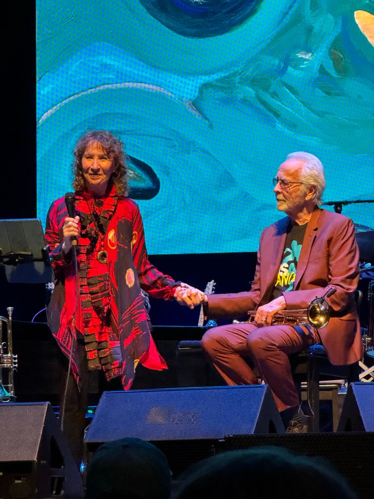 Herb Alpert sitting on a chair on stage holding the hand of his wife Lani Hall as she prepares to sing a mesley of Sergio Mendes & Brasil ‘66 songs.