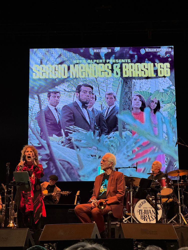 Lani Hall standing on stage singing while Herb Alpert listens from his seat next to her. The cover of the iconic album Herb Alpert Presents Sergio Mendes & Brasil ‘66 is on the big screen behind them.