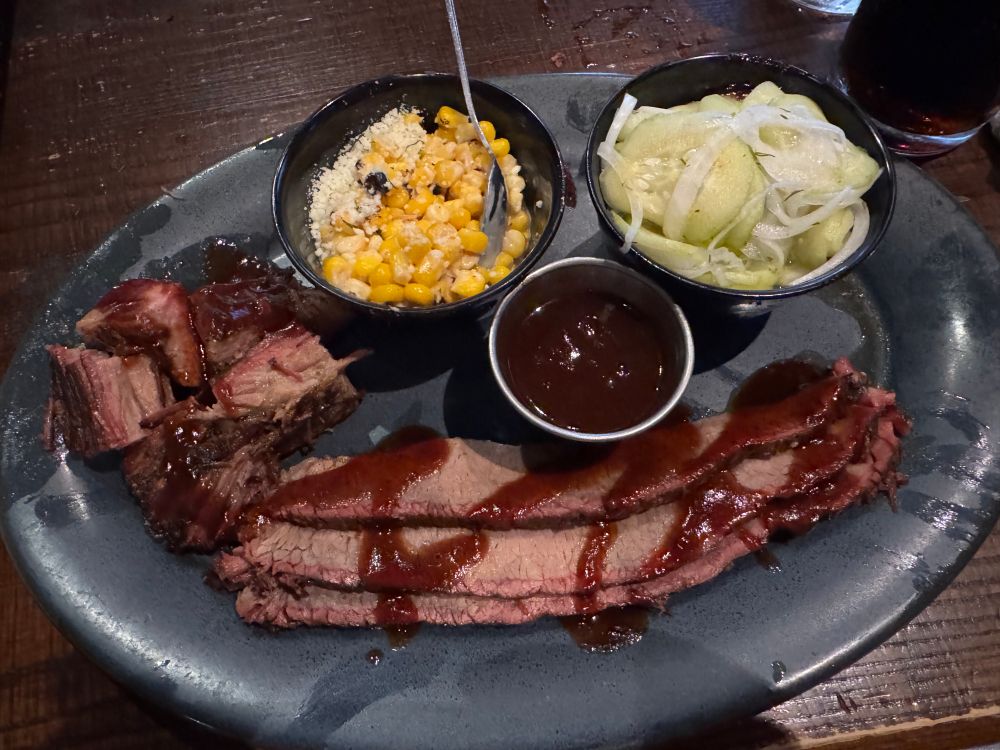 Platter with smoked brisket slices, several burnt ends, bbq saice, street corn, and cucumber-onion salad. 