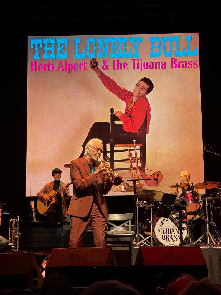 Herb Alpert on stage playing a trunpet with the Tijuana Brass behind him and the Lonely Bull album cover projected on the screen behind him.