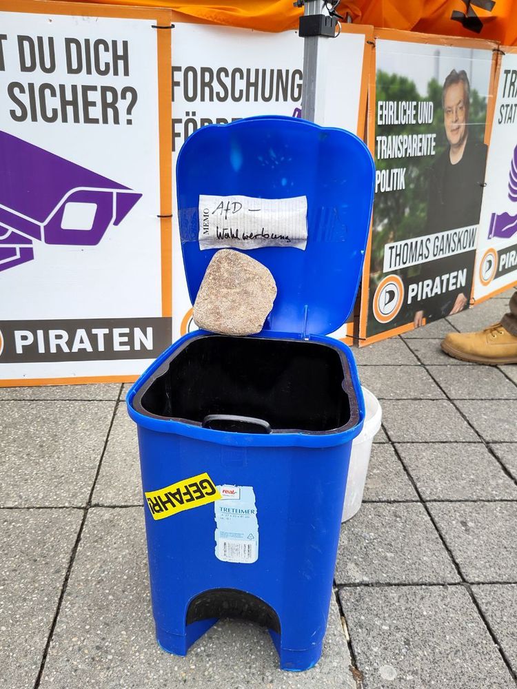 Eine blaue Mülltonne mit geöffnetem Deckel der durch einen Stein offen gehalten wird. Im Deckel ein Zettel mit der Aufschrift AfD Wahlwerbung.
Ein gelber Aufkleber mit schwarzer Schrift klebt vorne noch drauf. Auf dem steht in Großbuchstaben "Gefahr".
Im Hintergrund sind Plakate der Piraten aufgestellt.