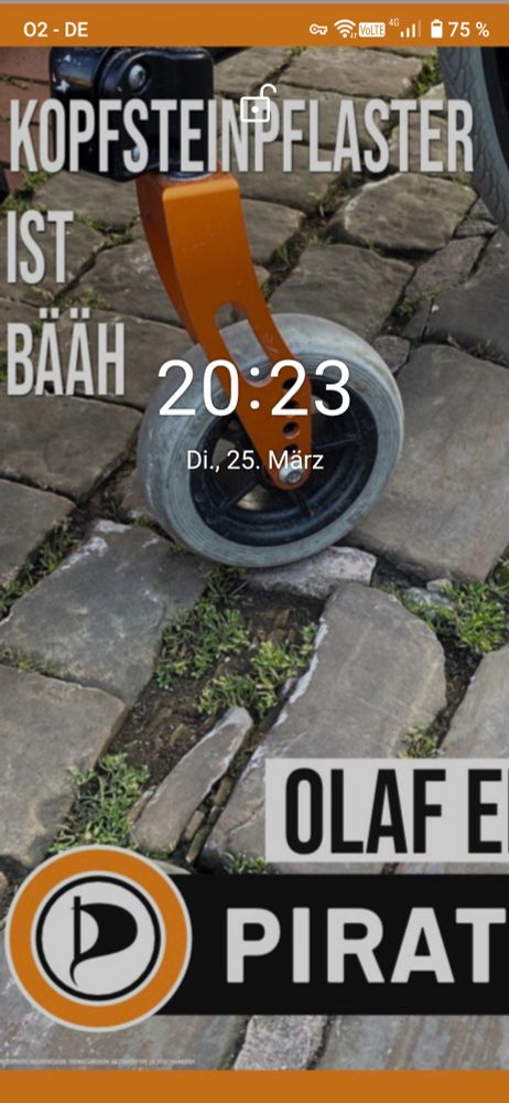 Kopfsteinpflaster mit tiefem Loch dahinter eine kleine Lenkrolle von einem Rollstuhl welche gleich da rein rutscht. Text oben links: Kopfsteinpflaster ist bääh. Unten rechts steht Olaf E. Darunter das Piratenpartei Logo und daneben das Wort Pirat. In der Mitte die Uhrzeit und das Datum.