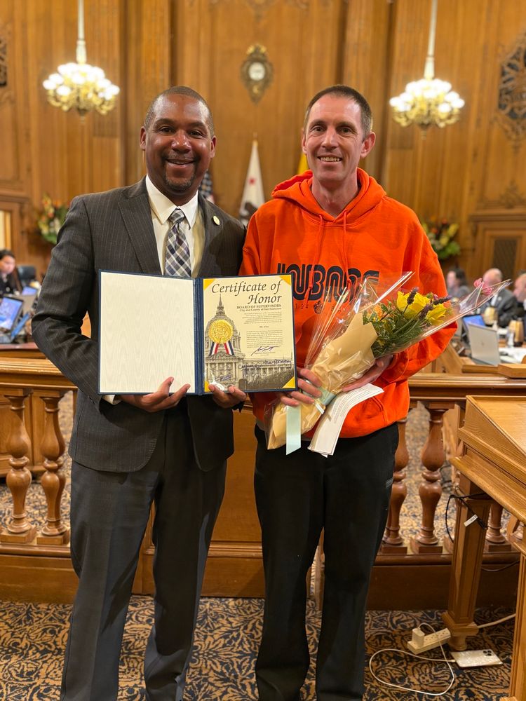 Supervisor Walton holding a certificate standing next to Mc Allen in an orange hoodie, holding flowers. They are standing in the San Francisco City Hall legislative chamber.