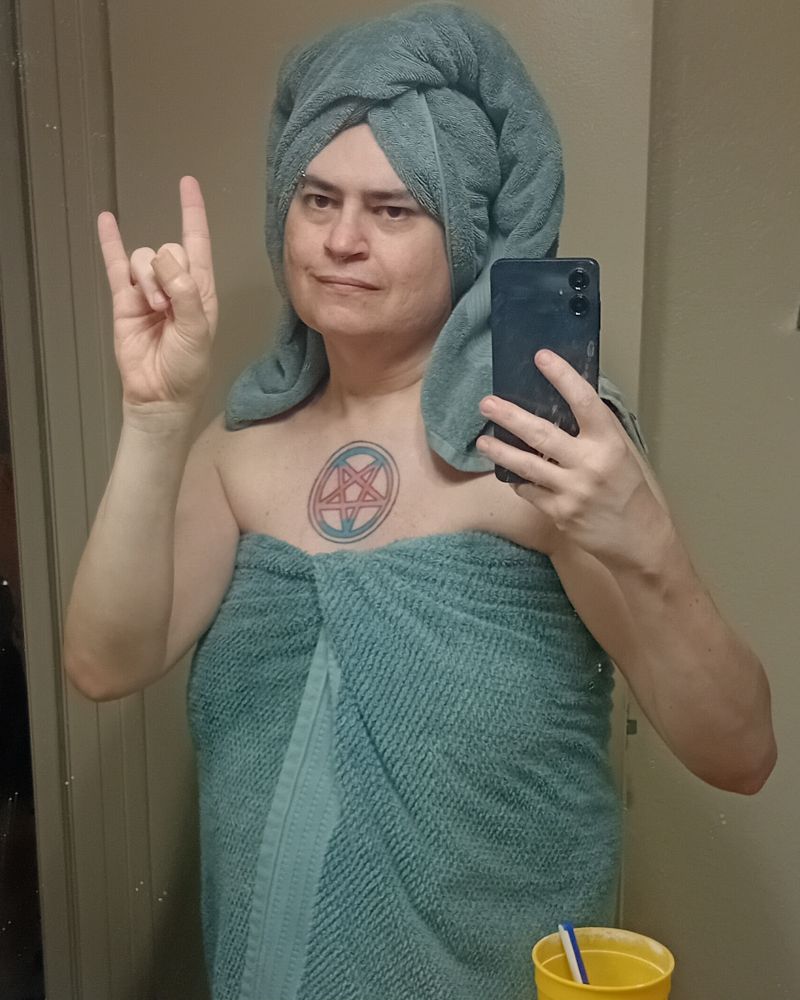 Pretty trans girl in a green post-bath towel and hair wrap giving devil horns to the mirror as she takes a selfie and oooh what's that it's a kickass pentagram in trans pride colors tattooed in the center of her breastbone let's goooooo