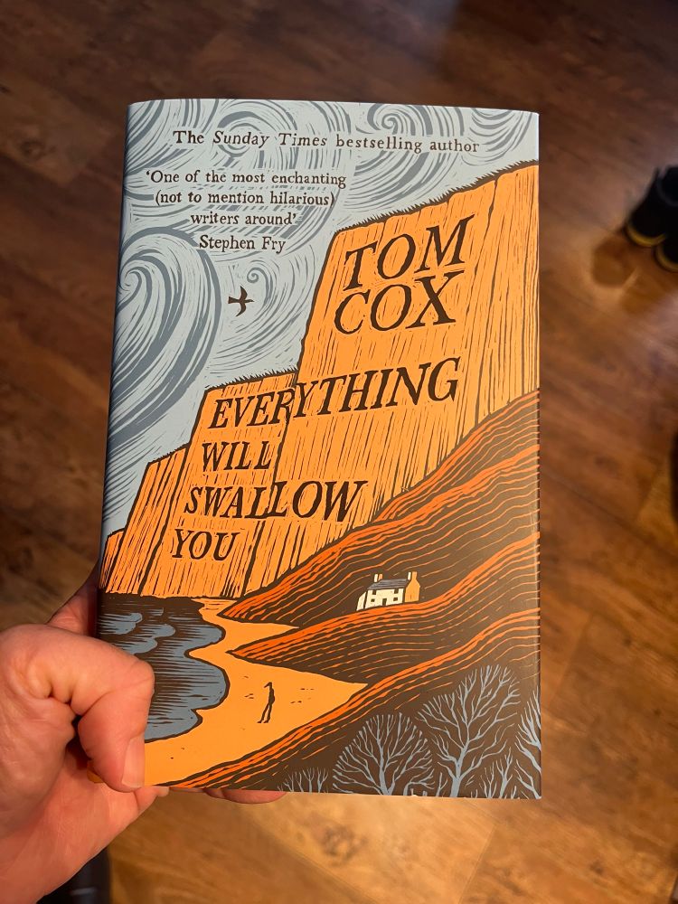 A hand holding out a hardback copy of Everything Will Swallow You by Tom Cox.