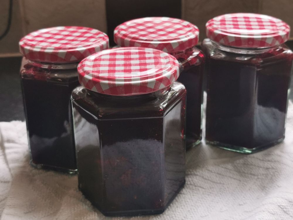  Jam Jars with a dark homemade blackberry jam, clear hexagonal jars with a red and white gingham (checkerboard) pattern. 