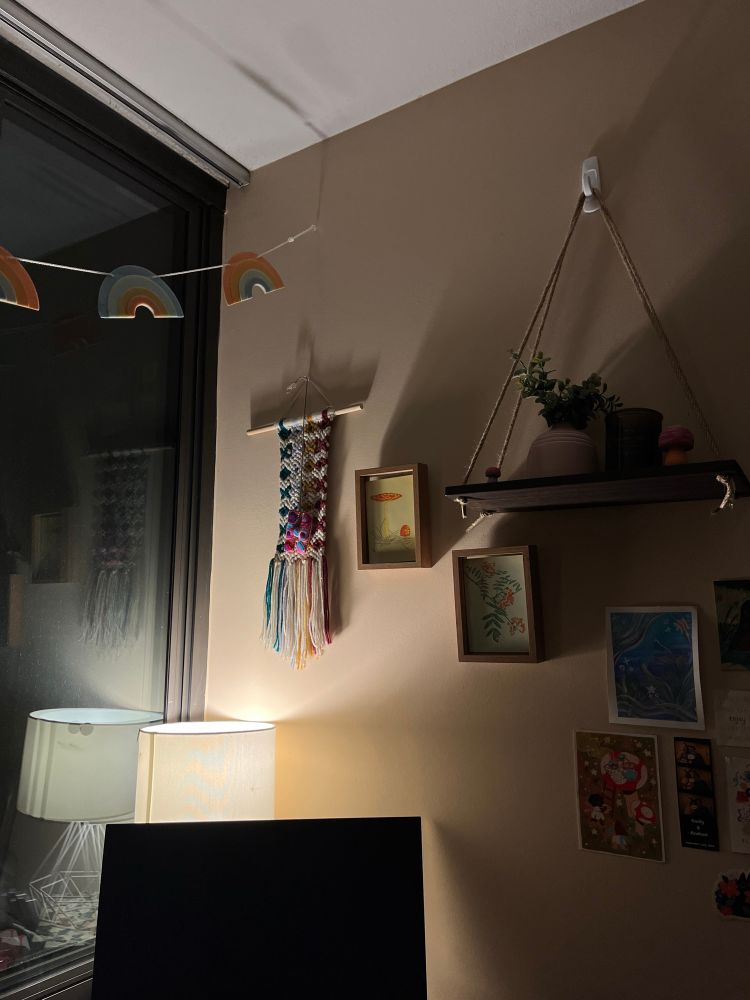 a table lamp illuminates the corner of a room, a computer monitor sits on top of a desk and wall art hangs behind it. There is a shelf with a plant, a macrame wall hanging, and a few postcards among the wall art. A string of rainbows on a garland hangs across the window to the left of the computer screen. 
