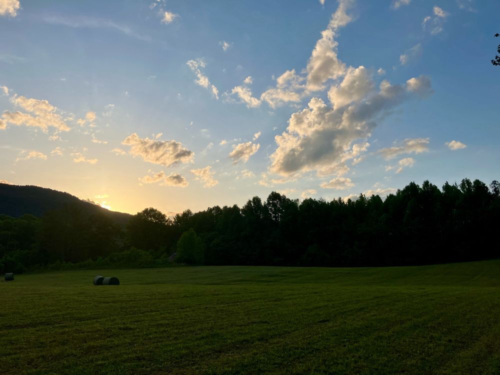 Sunset behind the mountains, with hay bale field in front of it. Fun, poofy clouds above in a pale blue sky.