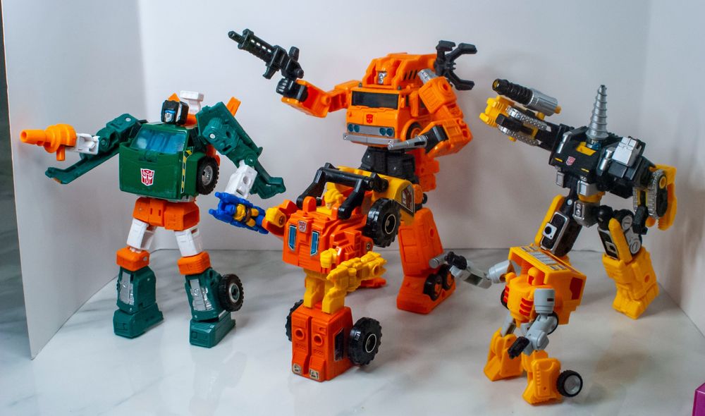 A group of construction-themed Autobots, in robot mode, in battle poses. Left to right: Earthrise Hoist, G1 Scoop, Earthrise Grapple, iGear Shafter, and Generations Selects Zetar.
