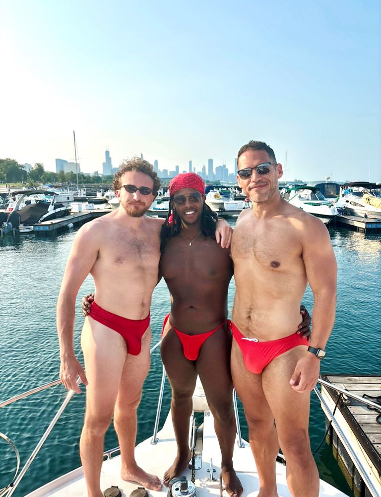 3 guys, standing next to each other on a boat, all wearing red swimwear, with the Chicago skyline in the background. 