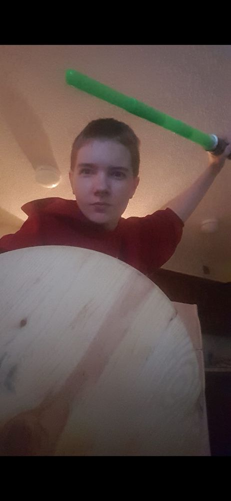 Photo of Hayden, in this he's wielding a green lightsaber over his head, poised to attack, while his right arm holds a circular wooden shield, unpainted. He's wearing a red hoodie and has very short brown hair. He's got an intense expression staring down the camera. End ID.
