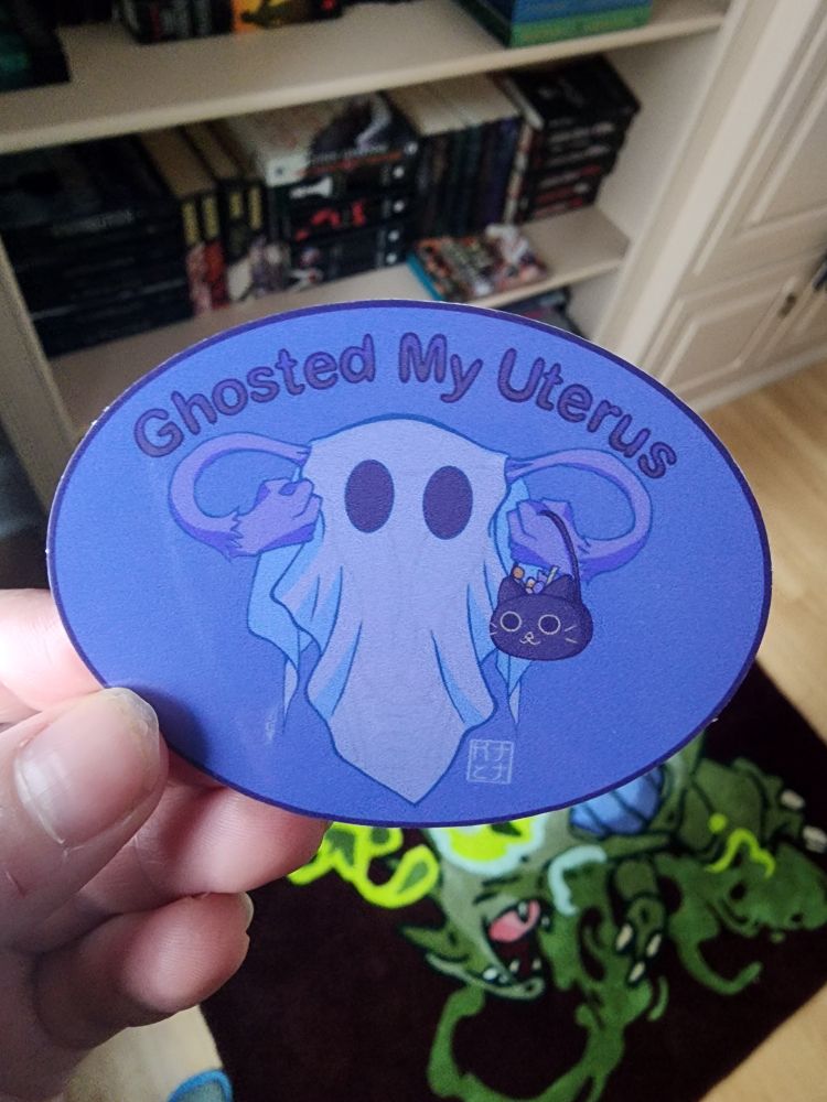 A sticker of a uterus dressed up like a ghost with a Halloween bag in the shape of a cat. It says, "Ghosted My Uterus"