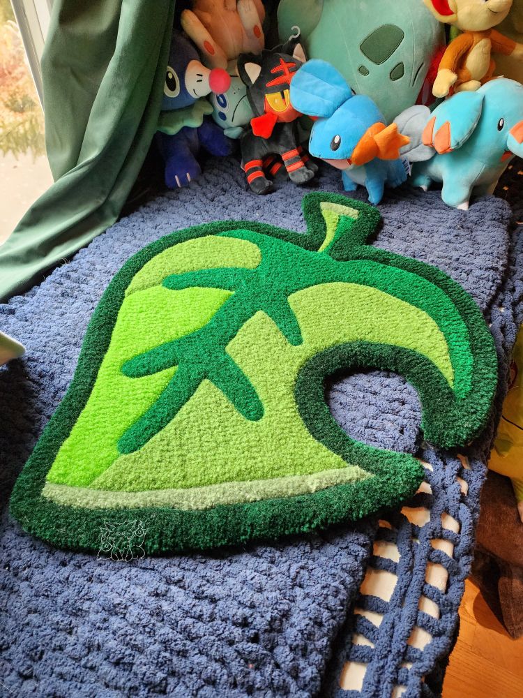 A handmade tufted leaf with various shades of green with a background of a window sill with many plush